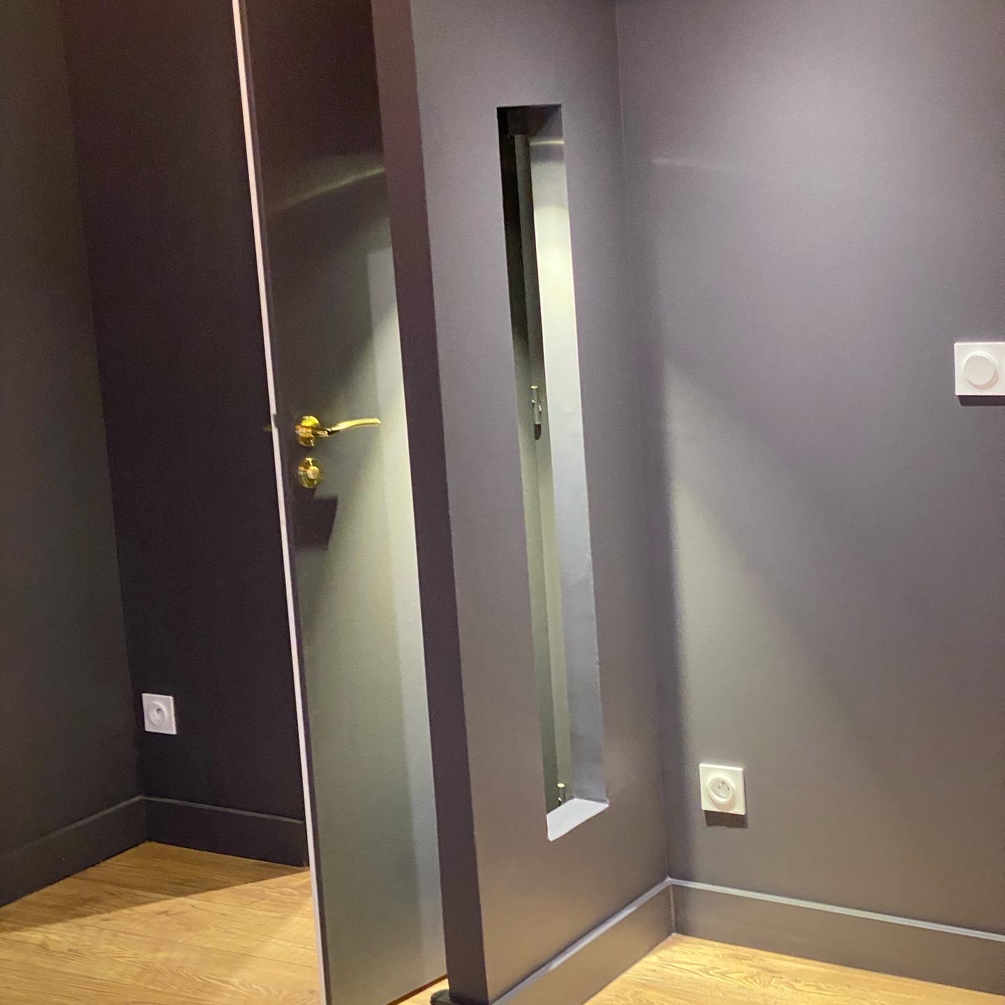 Cloison peinte gris anthracite avec ouverture verticale étroite, porte assortie à poignée dorée et sol stratifié effet chêne