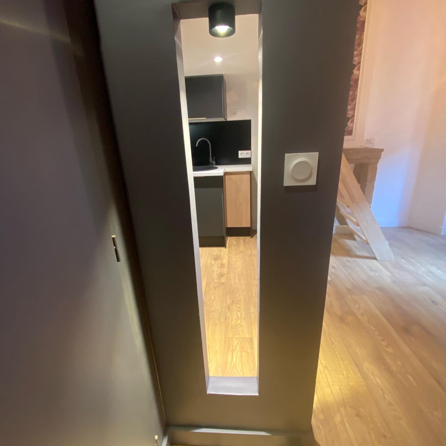 Ouverture verticale dans une cloison anthracite donnant sur une kitchenette noire et un sol stratifié effet chêne