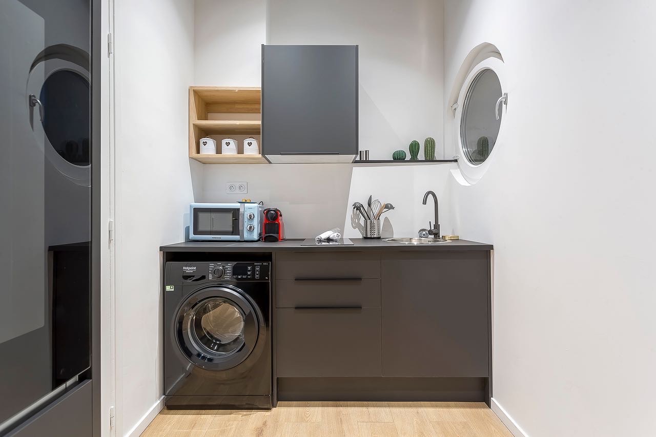 Kitchenette grise avec lave-linge sous plan, étagère en bois et fenêtre ronde type hublot intégrée dans un mur blanc